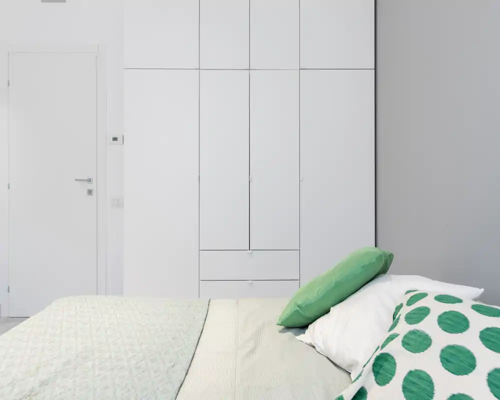 Bright, minimalist bedroom with a visible double bed in the foreground, white built-in wardrobe and green-accented pillows for a fresh, modern look.