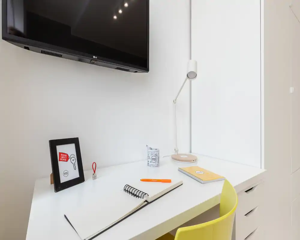 Espacio de trabajo luminoso y minimalista en la zona de estar con escritorio blanco, lámpara, TV en la pared y silla amarilla — moderno y ordenado para estancias cortas.