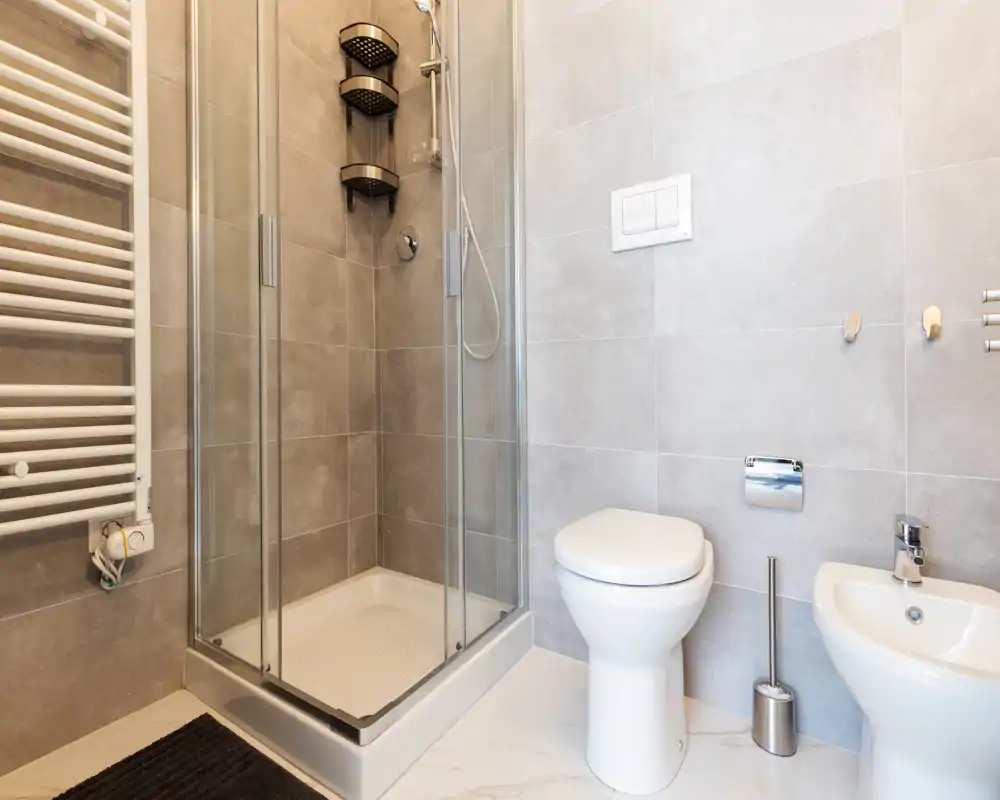 Baño moderno y limpio con mampara de ducha de vidrio, inodoro y bidé sobre azulejos gris claro.