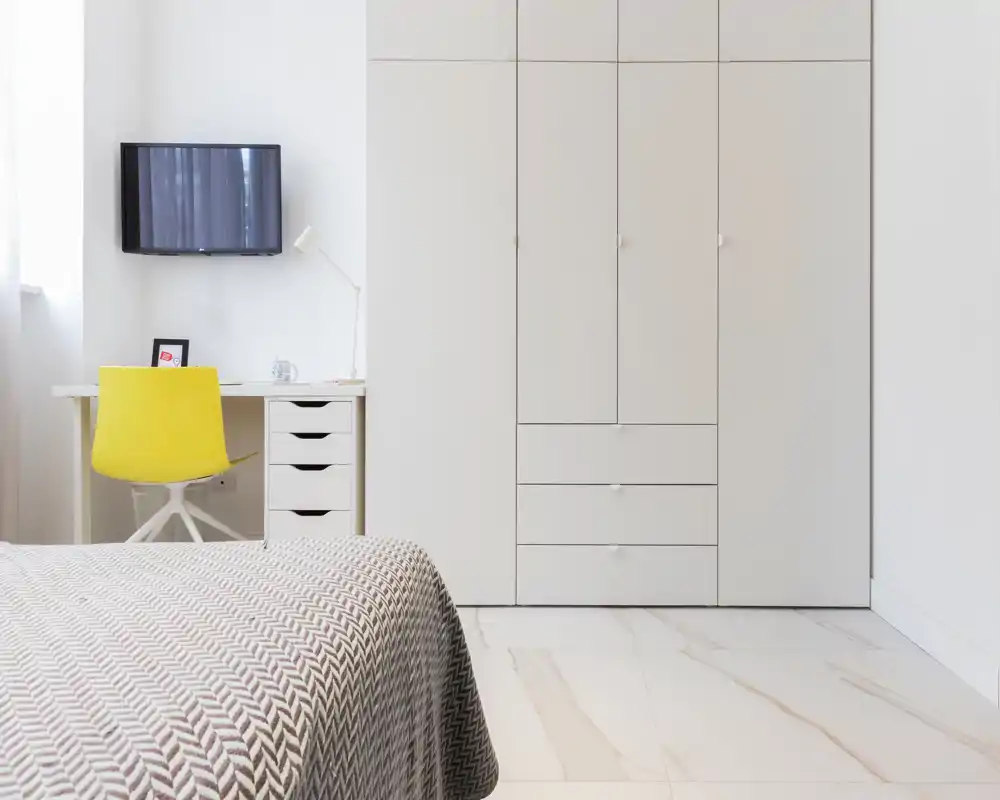 Dormitorio minimalista y luminoso con colcha de patrón, armario empotrado y un escritorio compacto con silla amarilla — moderno y bien iluminado.