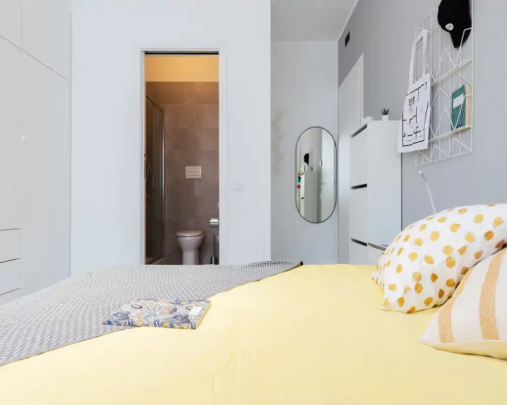 Dormitorio moderno y luminoso con cama vestida en amarillo, cojines decorativos y baño en suite visible; decoración limpia y minimalista con buena luz natural.