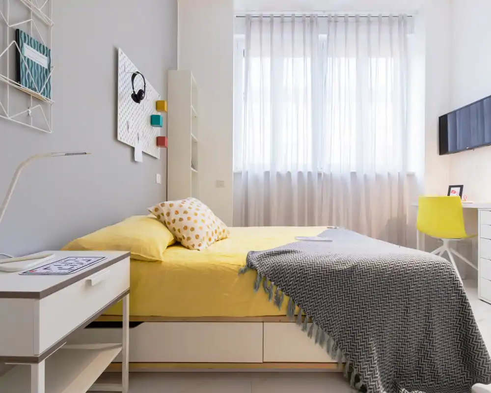 Dormitorio moderno y luminoso con colcha amarilla, cojín estampado y una manta gris acogedora; gran ventana que proporciona luz natural suave y una pequeña zona de escritorio/TV lateral.