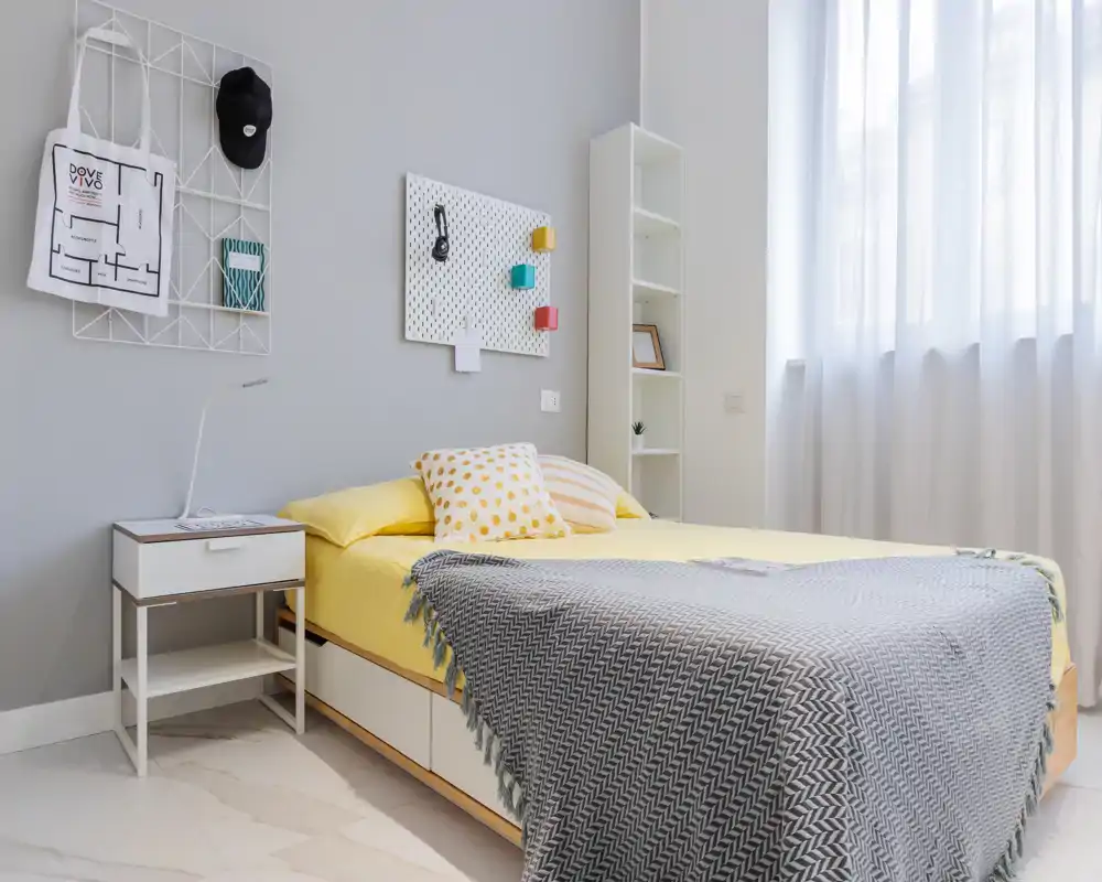 Dormitorio moderno y luminoso con cama doble bien hecha en ropa de cama amarilla, mesita con lámpara y estantería alta junto a la ventana — brillante y acogedor.