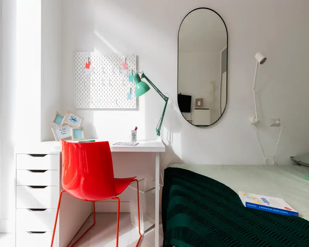 Angolo camera da letto luminoso e moderno con letto ben rifatto e una scrivania bianca compatta. Una sedia rossa e una lampada verde danno tocchi di colore; uno specchio ovale e decor minimal creano un look pulito e di stile.