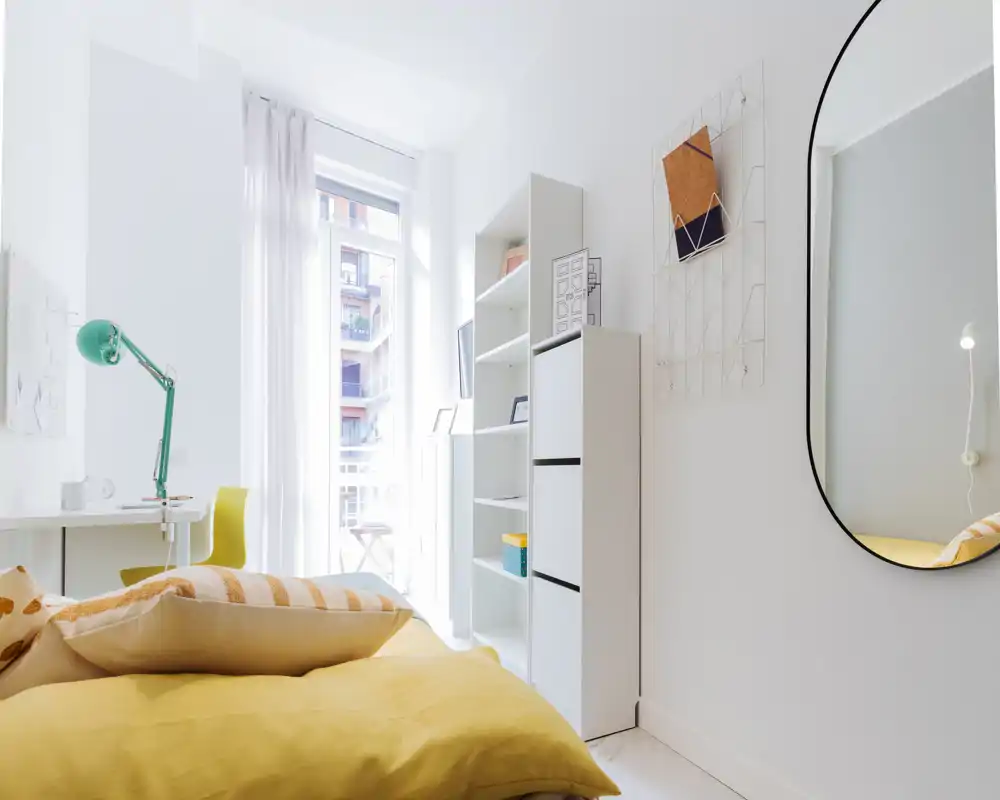 Bright, modern bedroom with a yellow bedspread, decorative cushions and a large window letting in abundant natural light. A white shelving unit, study desk with a green lamp and a wall mirror complete the stylish, airy space.