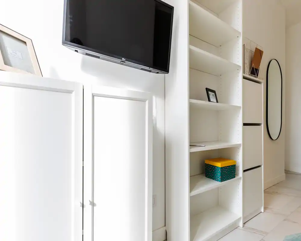 Bright, minimal living area corner with white built-in shelving and a wall-mounted TV; modern, clean styling with a decorative box and small framed items.
