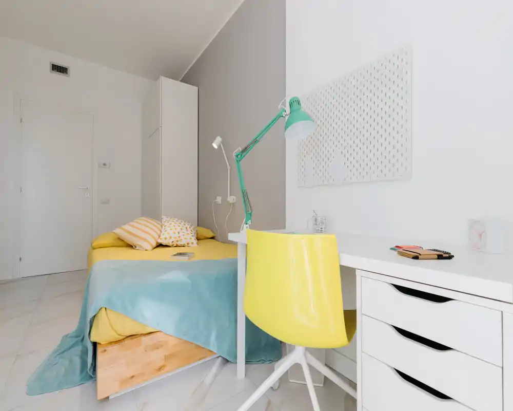 Bright, modern bedroom with a neatly made bed, pastel textiles and a small white desk with a yellow chair — ideal for both rest and study.