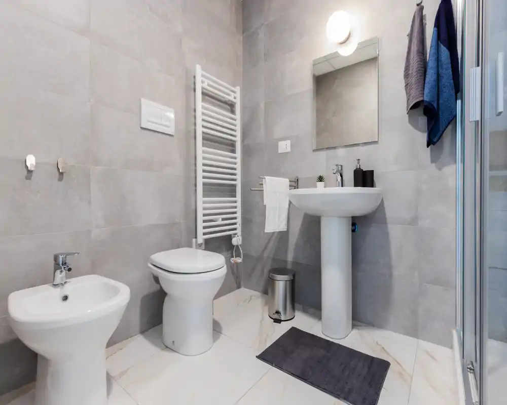 Baño moderno y bien iluminado con inodoro, bidé, lavabo de pedestal, espejo, radiador toallero y ducha visible en el borde de la imagen. Alicatado neutro y suelo efecto mármol para un estilo limpio y minimalista.