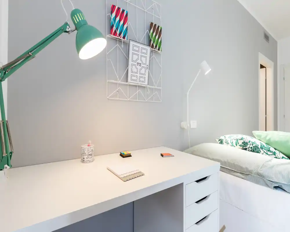 Rincón de dormitorio moderno y luminoso con un escritorio blanco ordenado y una cama cómoda con cojines estampados — ideal para trabajar y descansar.