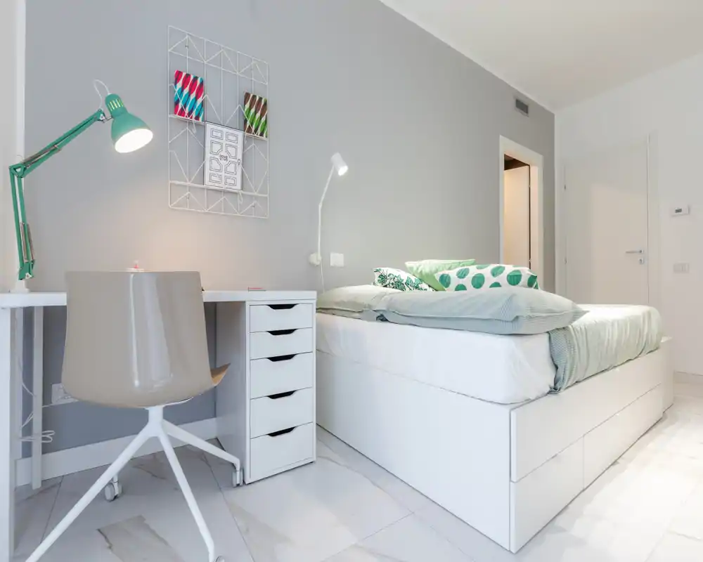 Dormitorio moderno y luminoso con cama plataforma blanca, cojines verdes y un escritorio compacto con lámpara — minimalista y bien iluminado, ideal para una persona.