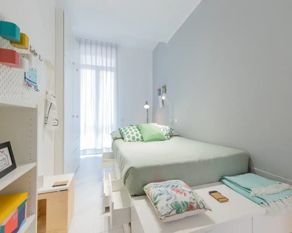 Dormitorio moderno y luminoso con cama doble en plataforma bien hecha, cojines decorativos y luz natural que entra por una ventana alta con cortina traslúcida.