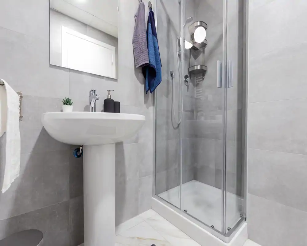 Baño moderno y compacto con ducha acristalada, lavabo con pedestal, espejo y azulejos grises neutras — luminoso, limpio y funcional.