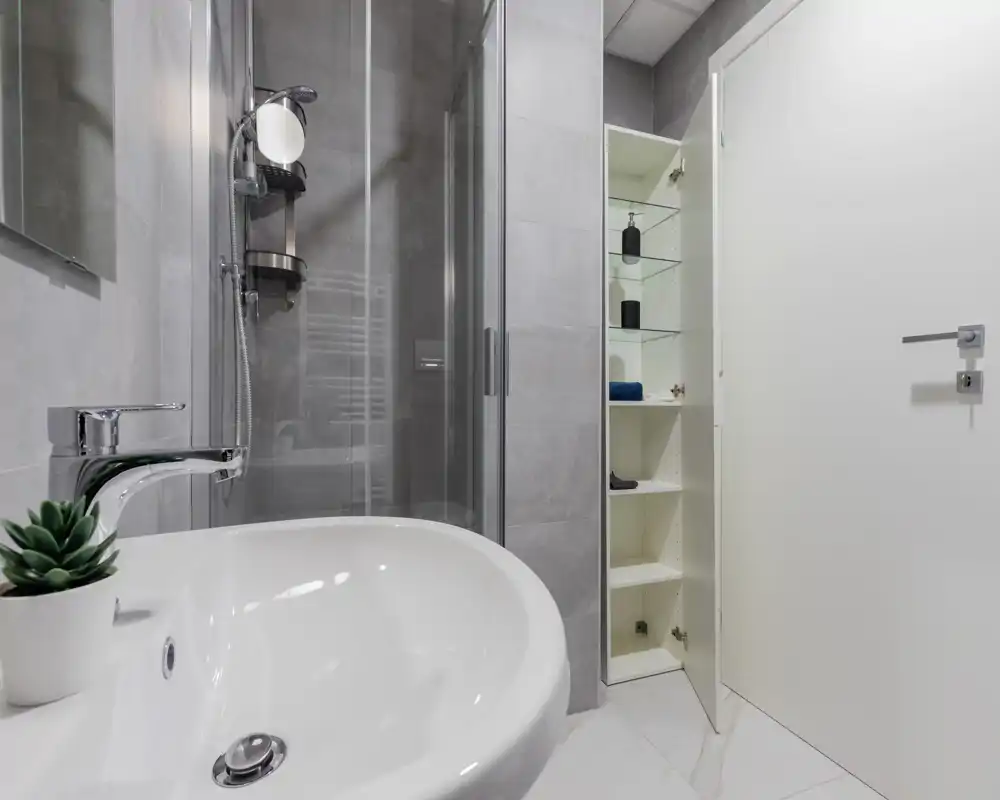 Baño moderno y compacto con lavabo blanco, grifo cromado y mampara de ducha de cristal. Estantes empotrados junto a la puerta ofrecen almacenamiento y los azulejos grises neutros crean un aspecto limpio y contemporáneo.