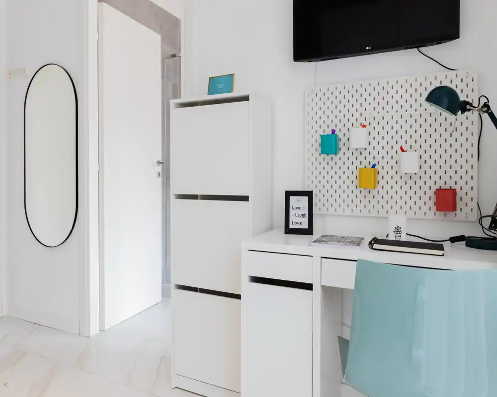 Rincón de estar/trabajo luminoso y minimalista con escritorio blanco, mueble de almacenamiento, organizadores en la pared y televisor — espacio moderno y limpio ideal para teletrabajo o descanso.