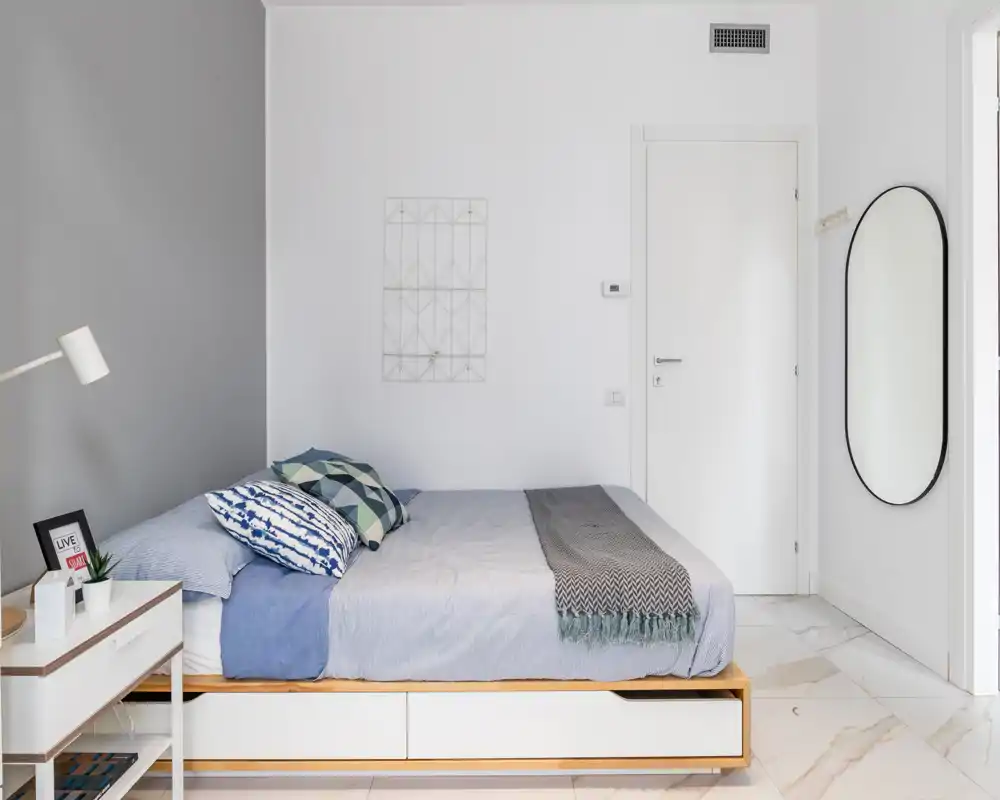 Dormitorio luminoso y minimalista con cama doble, cojines estampados y una manta. Paredes blancas, mesita de noche con lámpara y espejo ovalado junto a la puerta.