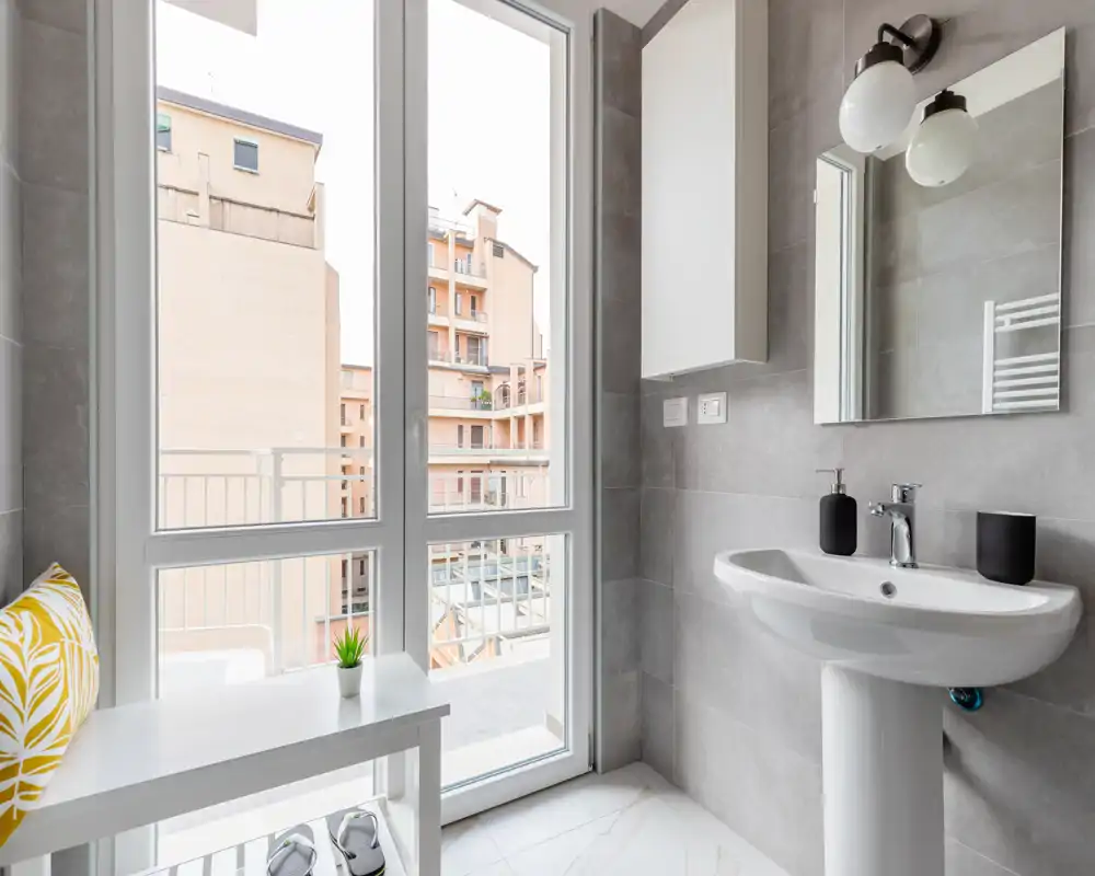Baño moderno y luminoso con lavabo de pie, espejo y gran ventana que da a un balcón; azulejos grises y accesorios minimalistas que ofrecen un aspecto limpio.