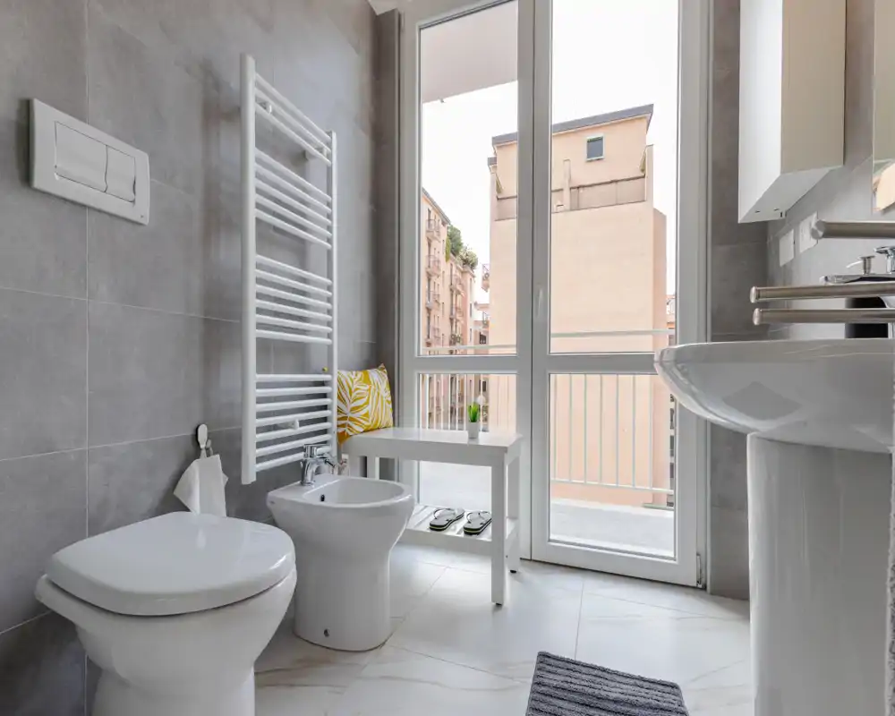 Baño moderno y luminoso con inodoro, bidé y lavabo de columna junto a una gran ventana que aporta luz natural. Acabados en azulejo y una pequeña mesa crean un aspecto fresco y minimalista.