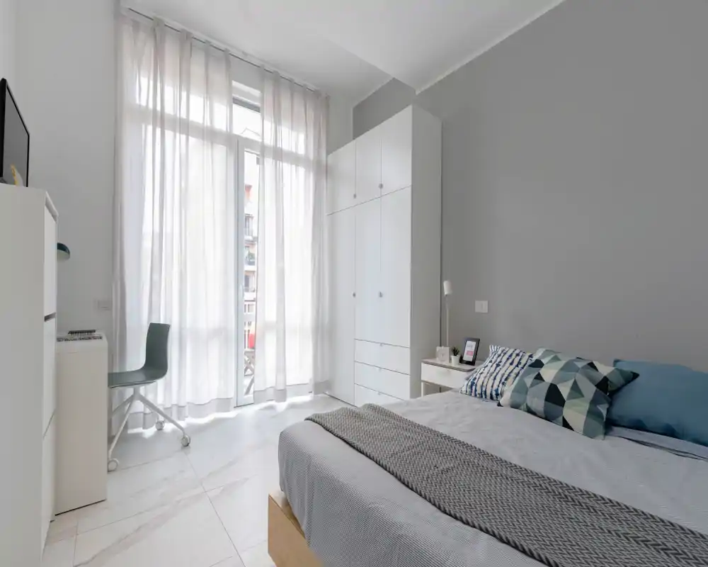 Dormitorio moderno y luminoso con cama doble, cojines geométricos y una gran ventana de suelo a techo que aporta mucha luz natural.