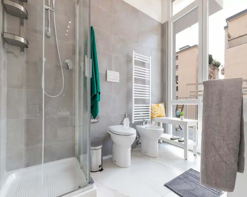 Baño moderno con ducha de vidrio, inodoro y bidé, amplia ventana que aporta luz natural y accesorios contemporáneos; suelo imitación mármol y paredes alicatadas neutras.