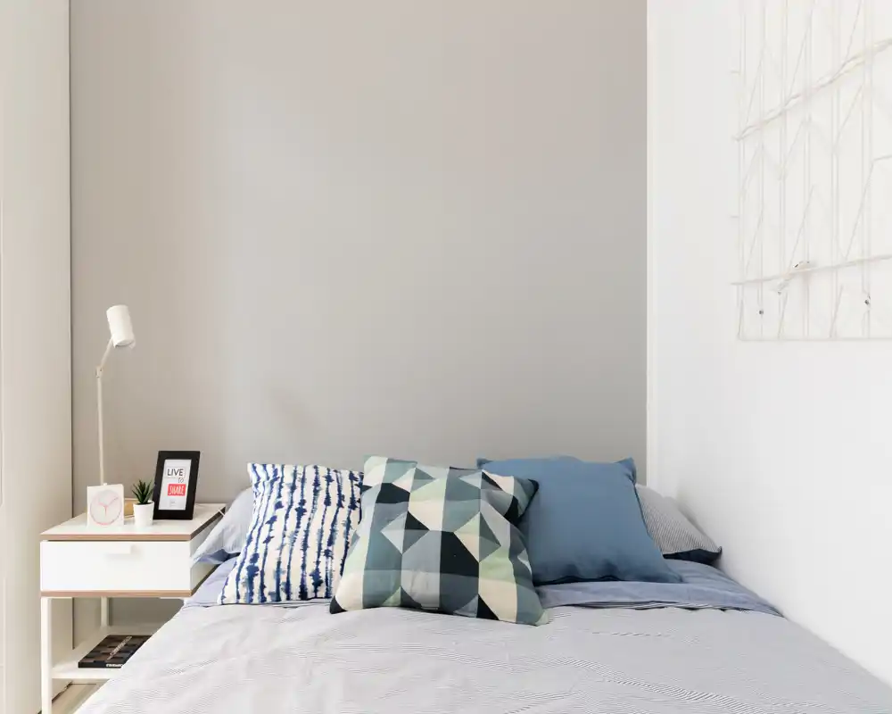 Dormitorio moderno y luminoso con cojines decorativos, cama ordenada y una mesita de noche minimalista con lámpara y accesorios — ideal para fotos de alquiler.