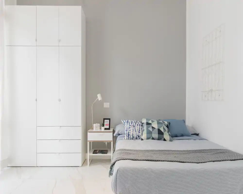 Dormitorio moderno y luminoso con cama doble, cojines decorativos y un armario alto blanco. Tonos neutros y mesita con lámpara crean un ambiente tranquilo y acogedor, perfecto para anuncios.