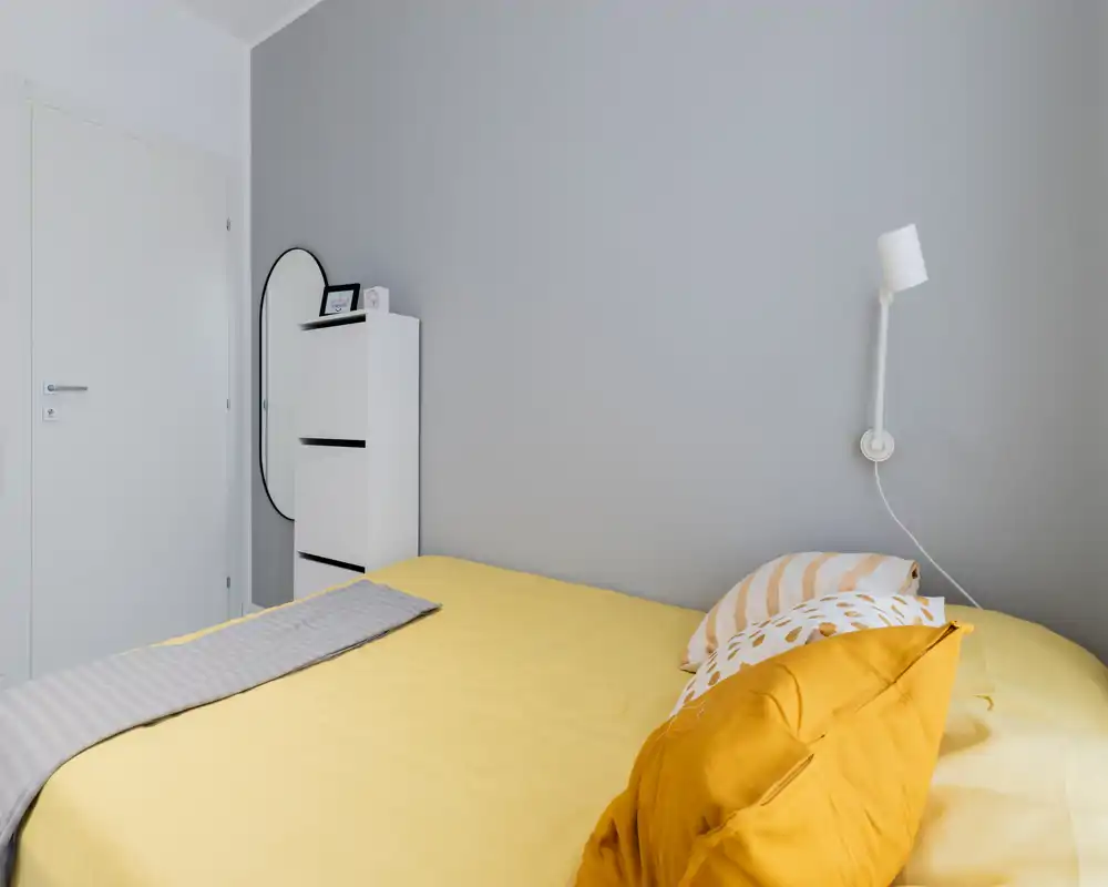 Dormitorio luminoso y minimalista con edredón amarillo, cojines en capas y una lámpara de pared simple: estilo moderno y limpio, ideal para el anuncio.