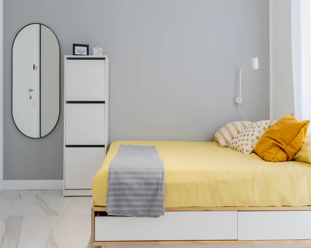Dormitorio moderno y luminoso con cama vestida en amarillo, cojines decorativos y un espejo de cuerpo entero, decorado con un estilo limpio y minimalista.