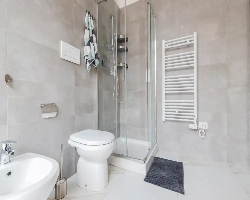 Baño moderno y luminoso con cabina de ducha de vidrio, inodoro, bidé y toallero eléctrico en la pared; azulejos grises y suelo efecto mármol para una apariencia limpia y contemporánea.