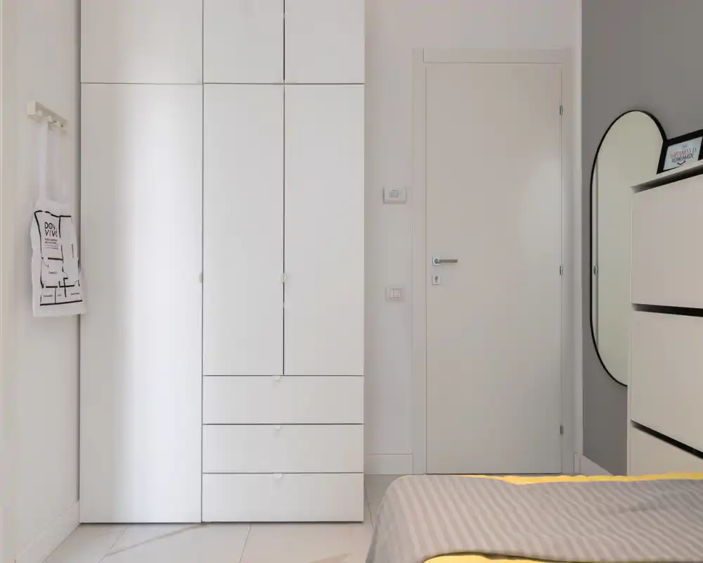 Rincón de dormitorio luminoso y minimalista con armario empotrado blanco, puerta cerrada y vista parcial de una cama bien hecha con ropa de cama sobria.