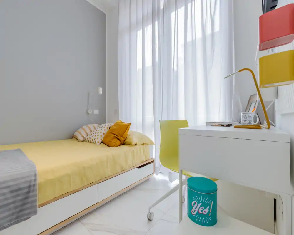 Habitación individual luminosa y moderna con colcha amarilla, cojines acogedores y un pequeño escritorio blanco junto a una gran ventana con cortina translúcida — ideal para estudiante o persona sola.
