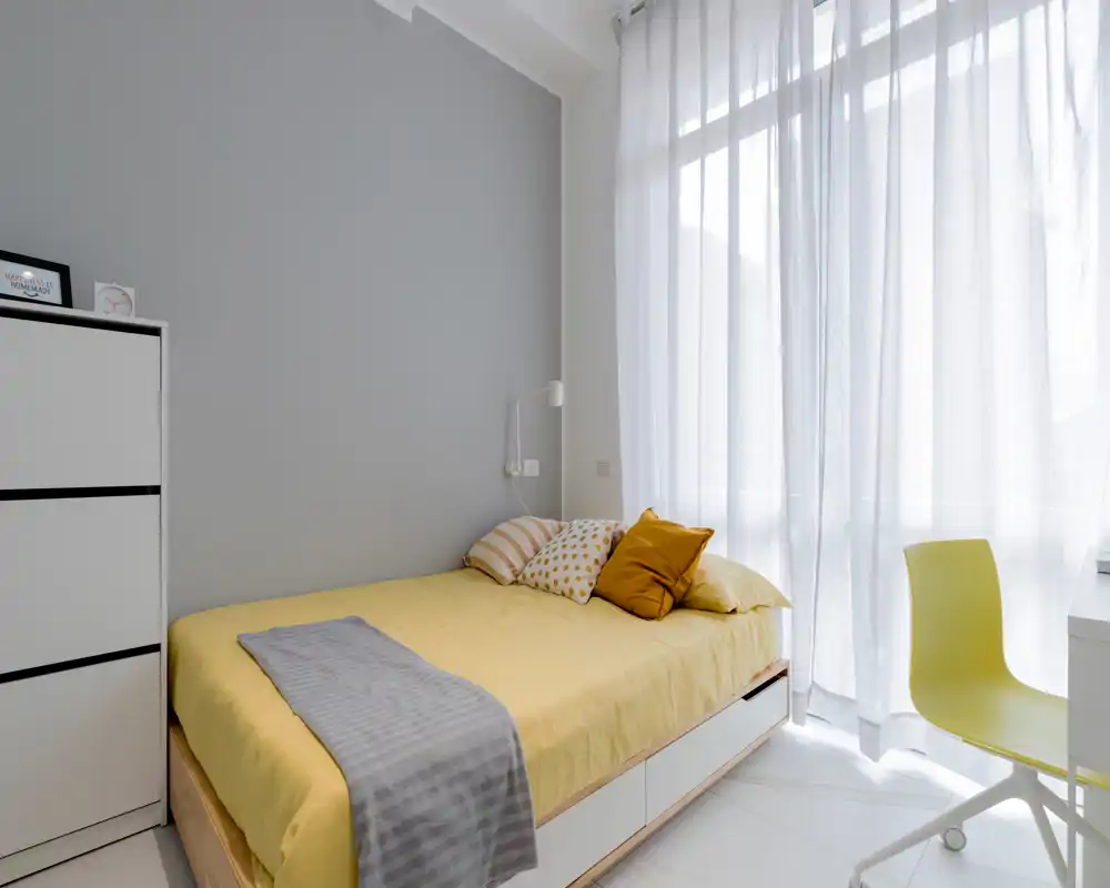 Dormitorio moderno y luminoso con cama doble bien hecha, ropa de cama amarilla, cojines decorativos y una gran ventana con cortinas finas que aportan luz natural.