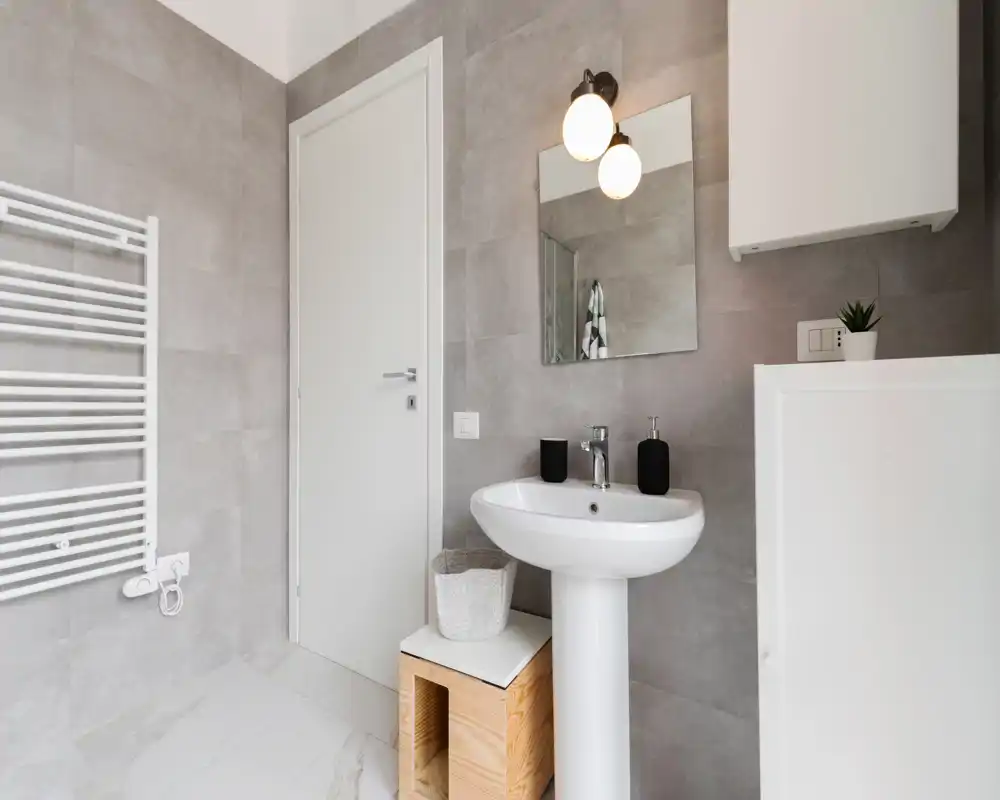 Baño moderno y luminoso con lavabo de pie, espejo, radiador toallero y azulejos grises neutras — diseño limpio y minimalista.