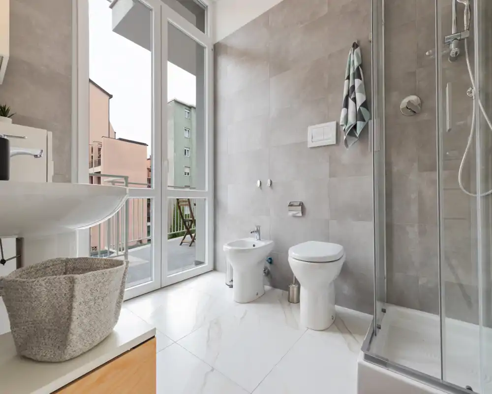 Baño moderno y luminoso con gran ventana, ducha, inodoro y bidé sobre suelo de efecto mármol.