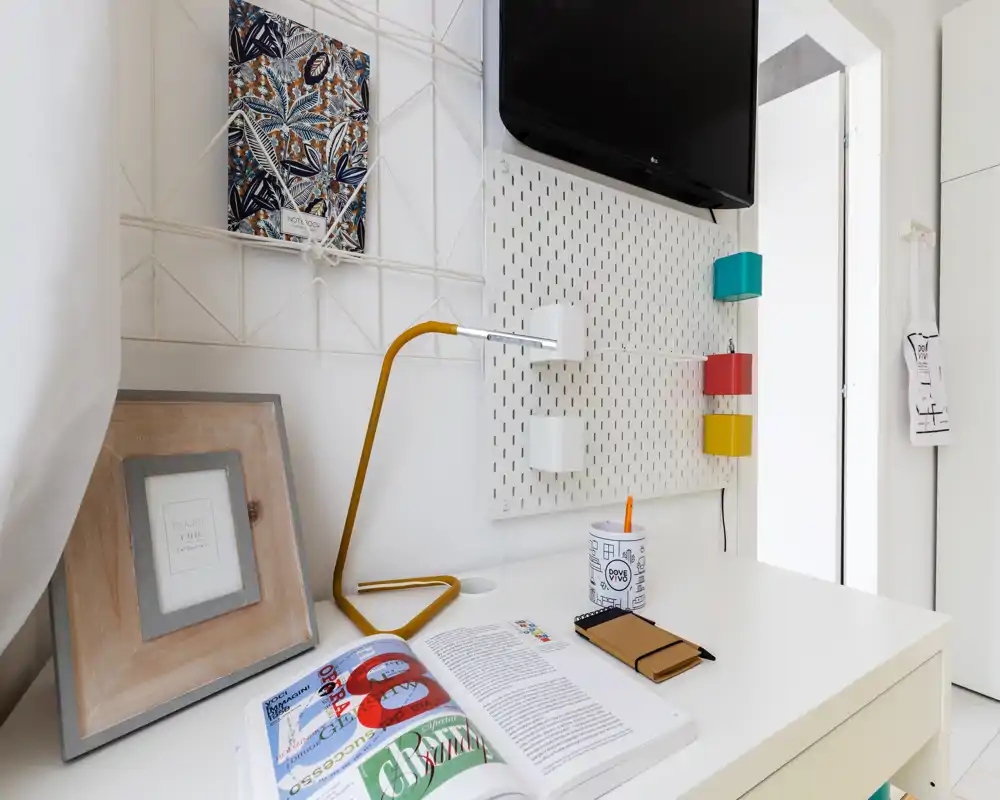 Espacio de trabajo luminoso en el dormitorio con escritorio blanco, marco decorativo, lámpara de mesa amarilla moderna y televisor en la pared — ideal para trabajar o leer.