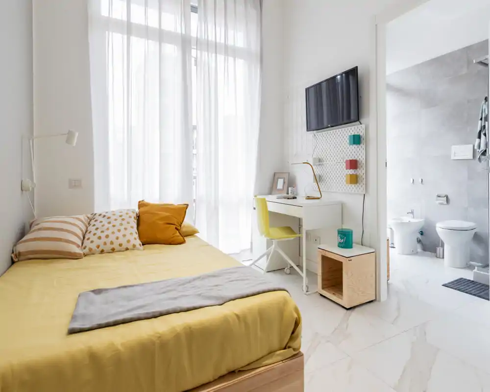 Dormitorio moderno y luminoso con cama bien hecha, ropa de cama amarilla, un escritorio y televisor mural; grandes cortinas translúcidas aportan mucha luz natural y se ve el baño en suite.