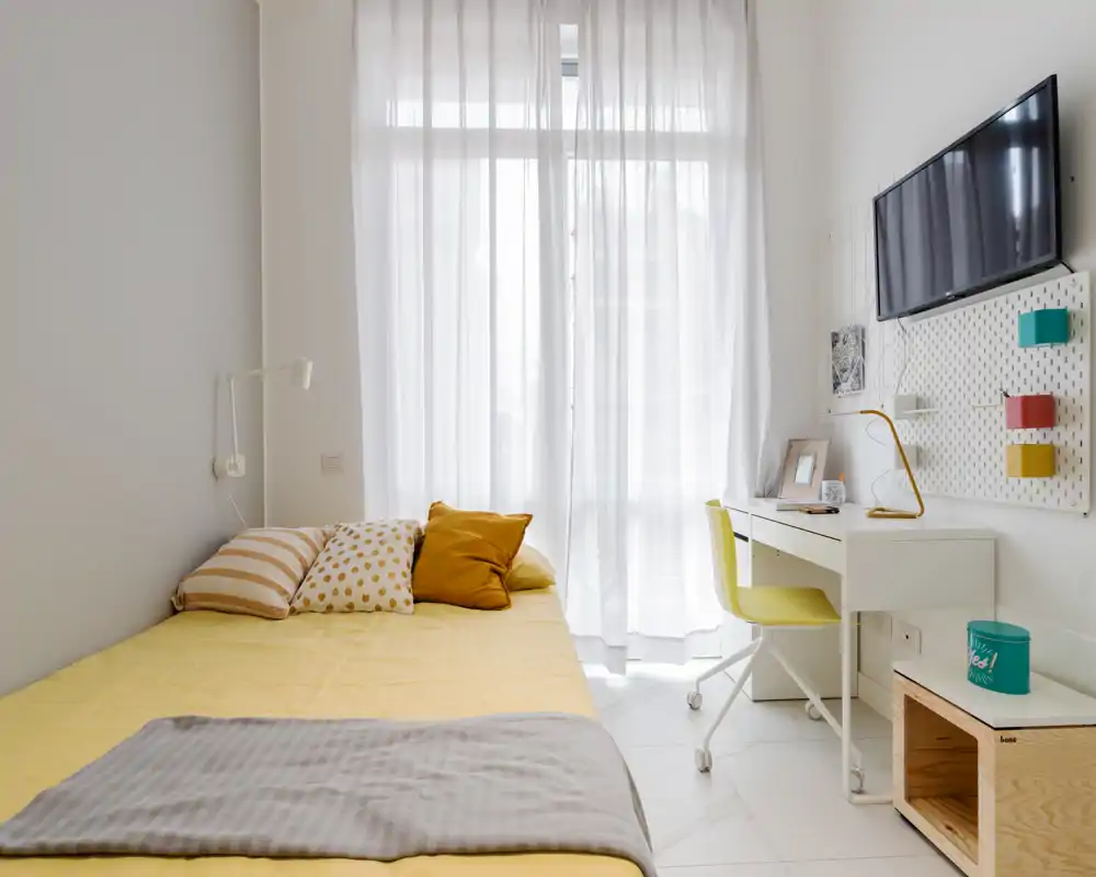 Dormitorio moderno y luminoso con cama individual bien hecha, ropa de cama amarilla cálida, cojines decorativos y un espacio de trabajo compacto con TV — ideal para una estancia acogedora.