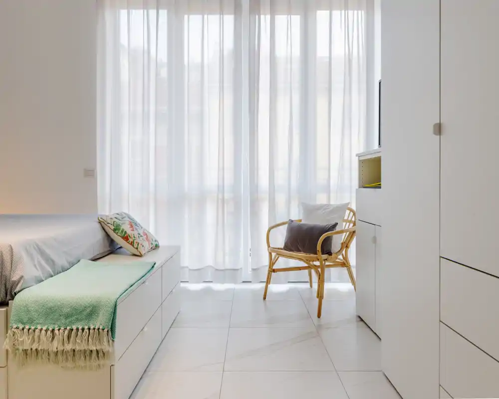 Dormitorio luminoso y minimalista con una cama amplia, textiles suaves y una silla de ratán junto a cortinas translúcidas — perfecto para anuncios que destaquen la luz natural y el estilo moderno y limpio.