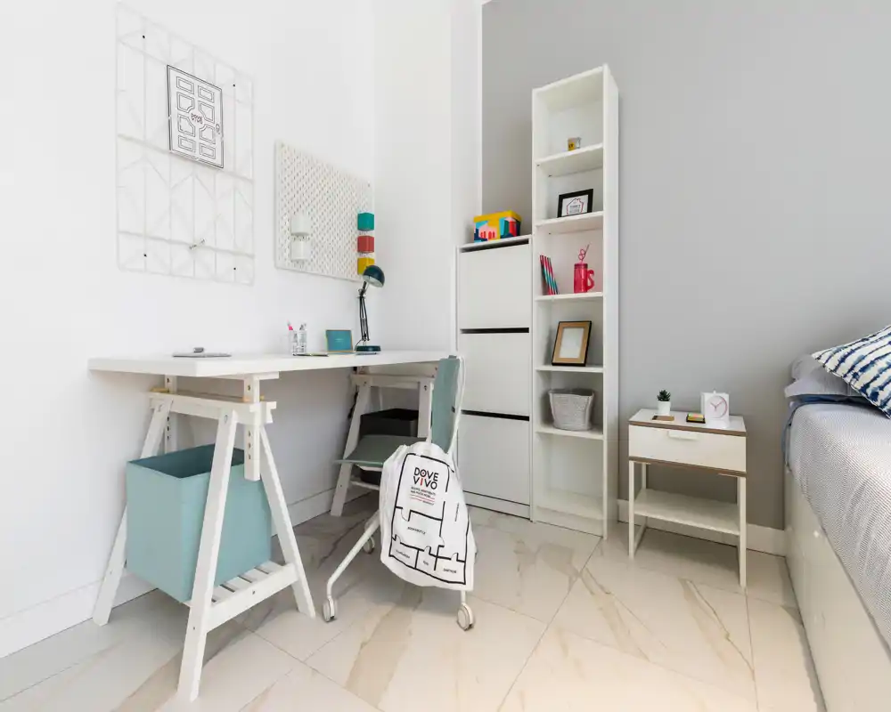 Rincón de dormitorio moderno y luminoso con escritorio y estantería: área de trabajo ordenada junto a una cama parcialmente visible con cojines a rayas, mesita elegante y suelos claros.