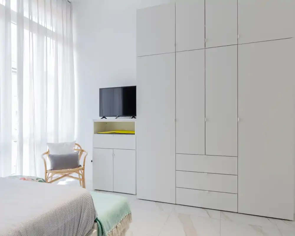 Dormitorio luminoso y minimalista con la cama en primer plano, grandes armarios empotrados, mueble para TV y una butaca de mimbre junto a una ventana con cortina traslúcida que aporta mucha luz natural.