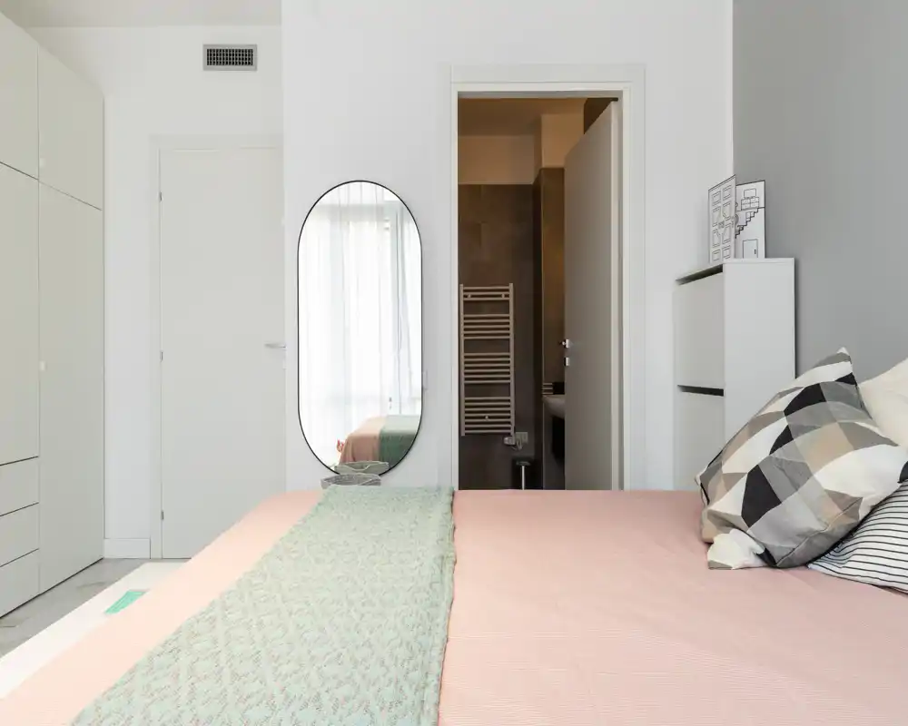 Dormitorio moderno y luminoso con cama bien hecha, cojines estampados y una manta verde; armario de gran tamaño y espejo en la pared que amplían el espacio.
