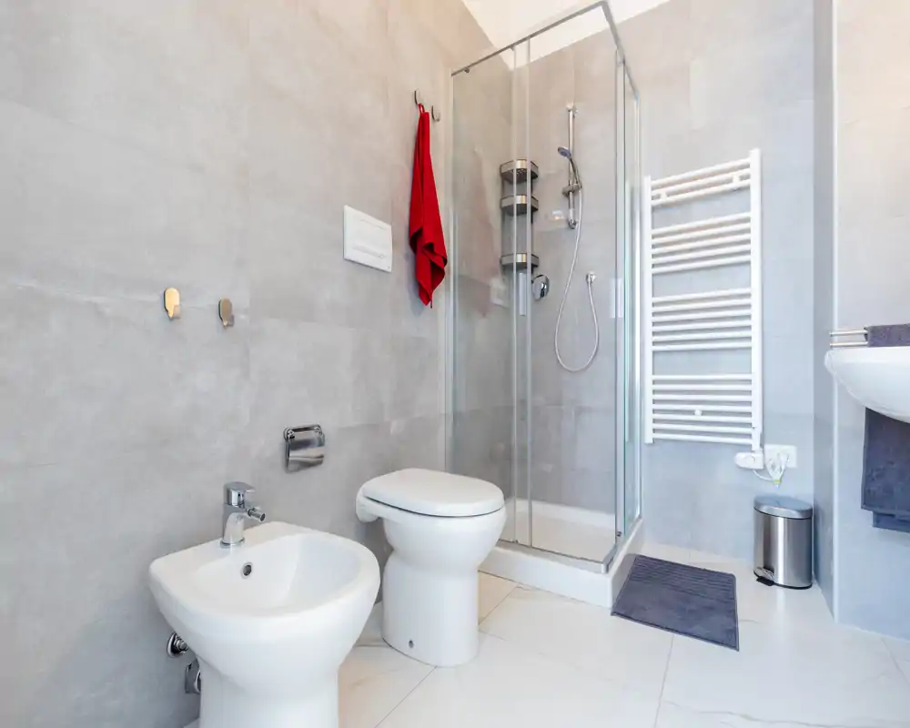 Baño moderno y luminoso con ducha de esquina de cristal, inodoro, bidé y radiador toallero mural. Azulejos neutros y accesorios contemporáneos ofrecen un aspecto limpio y minimalista.