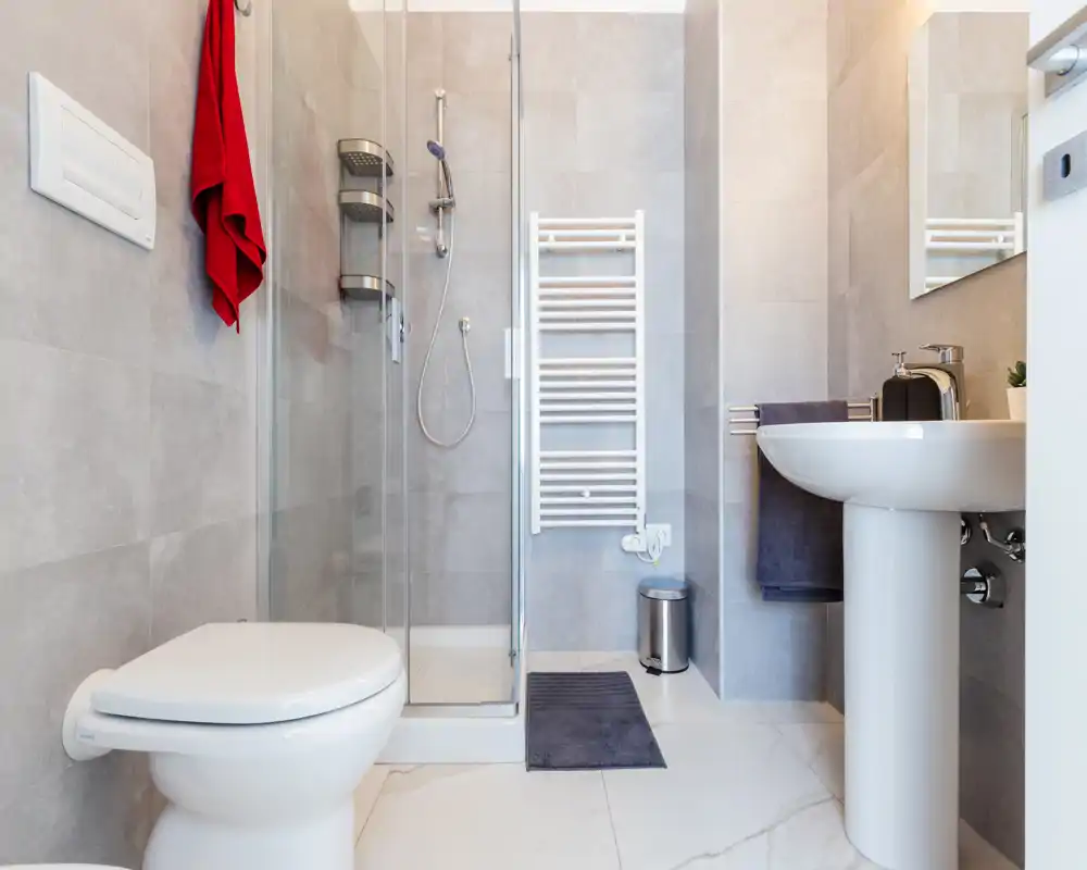 Baño moderno y luminoso con ducha de vidrio, lavabo de pie, inodoro mural y toallero calefactado. Baldosas grises neutras y accesorios mínimos ofrecen una apariencia limpia y contemporánea.