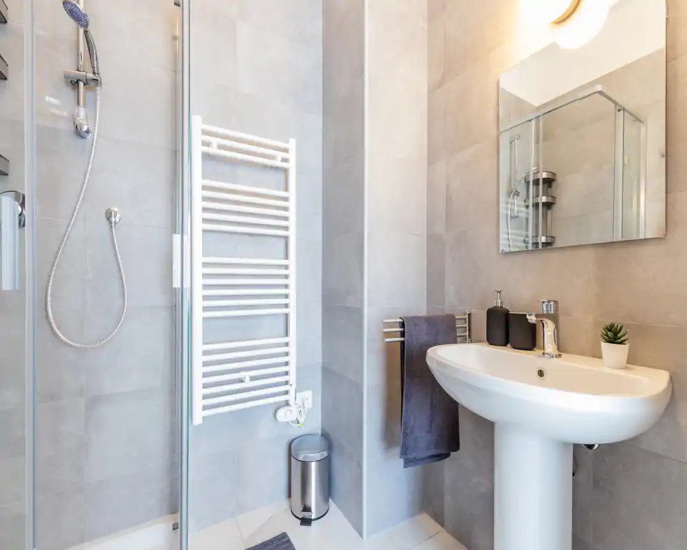 Baño moderno y limpio con lavabo de pedestal, espejo y ducha de cristal. Baldosas grises y accesorios ordenados crean un espacio funcional y luminoso.