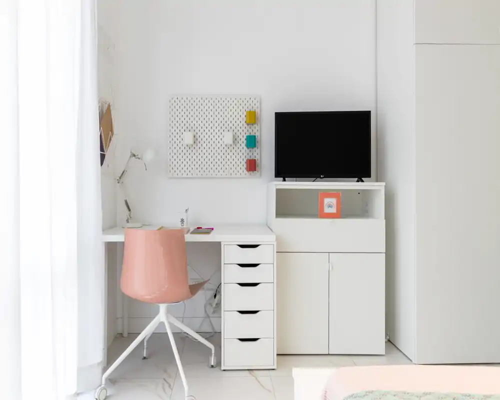 Rincón de dormitorio luminoso y minimalista con escritorio blanco, silla giratoria rosa y un pequeño mueble con TV; se aprecia parte de la cama en la esquina inferior derecha, indicando que es un dormitorio con zona de trabajo.