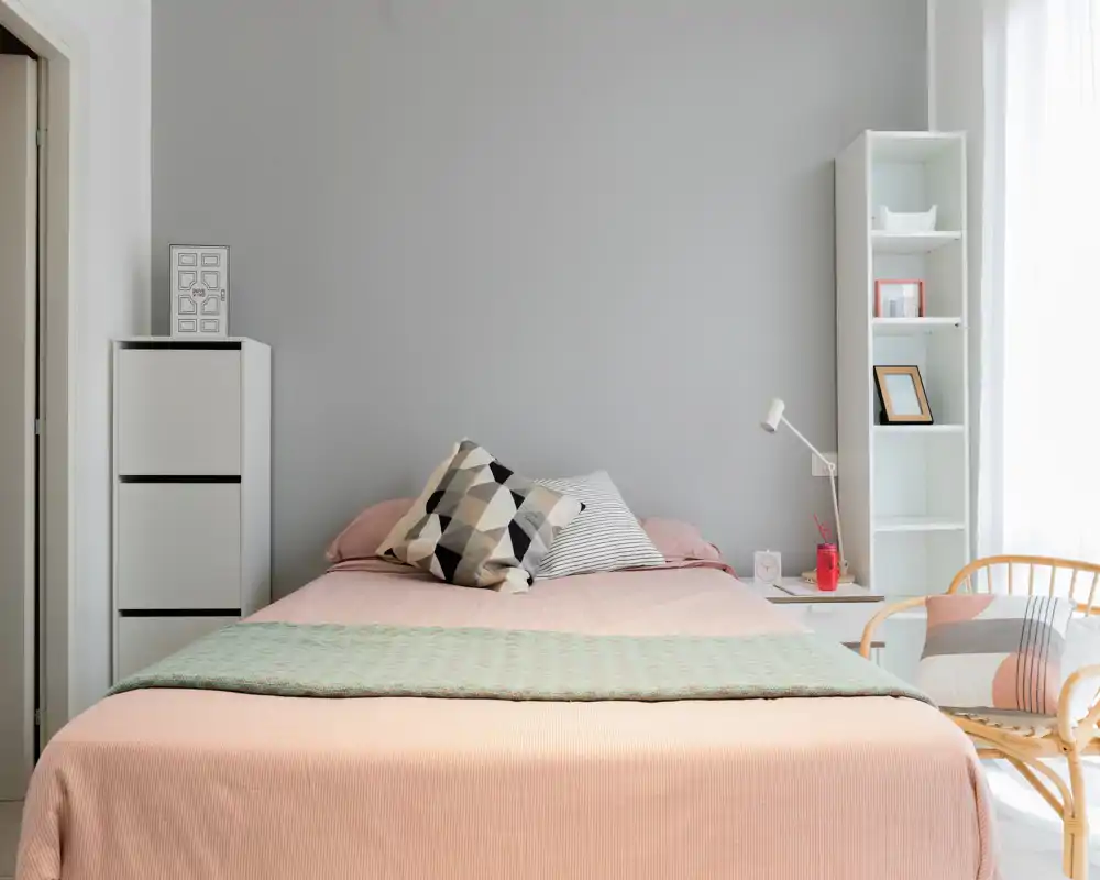 Dormitorio moderno y luminoso con una cama grande bien hecha, ropa de cama en tonos pastel y estanterías minimalistas — un espacio tranquilo y acogedor con luz natural.