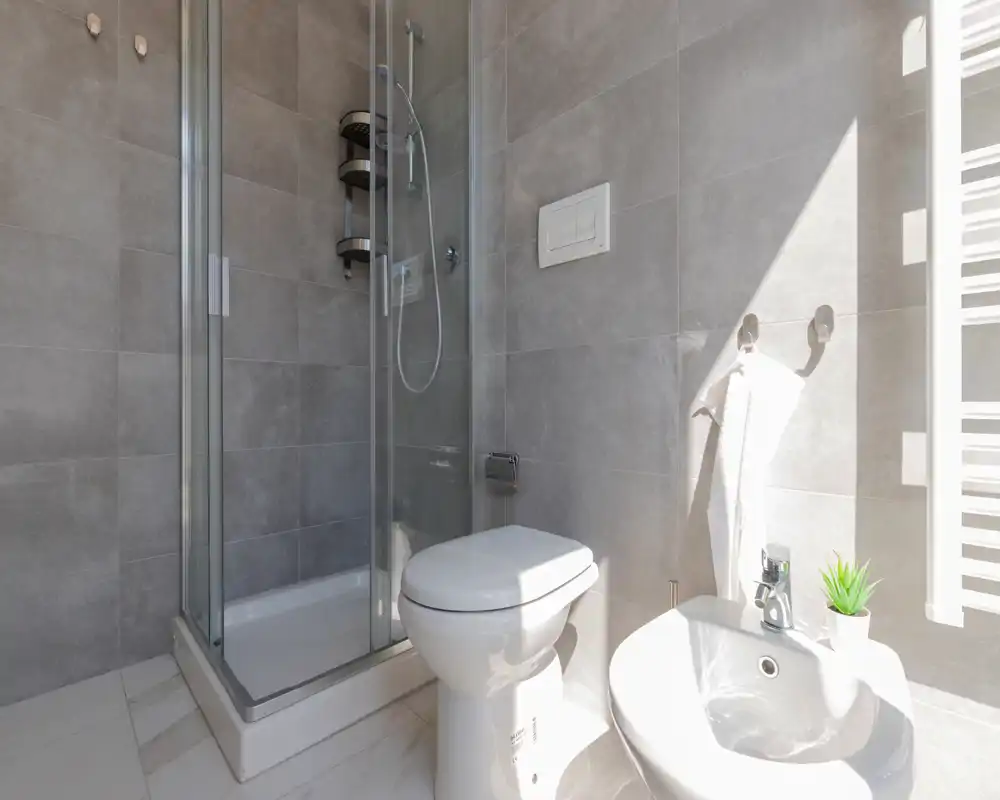 Bright, modern bathroom with a corner glass shower, wall-mounted toilet flush plate, bidet, and towel hooks; neutral grey tiles and marble-effect floor create a clean contemporary look.