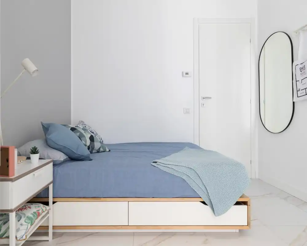 Bright minimalist bedroom with a low wooden bedframe, blue linens, decorative pillows and a knit throw; clean white walls and a standing mirror create an airy, modern feel.
