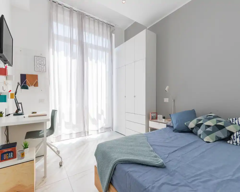 Bright, modern bedroom with a comfortable double bed, bedside table and large wardrobe. Natural light floods the room through sheer curtains, creating a fresh and airy atmosphere.