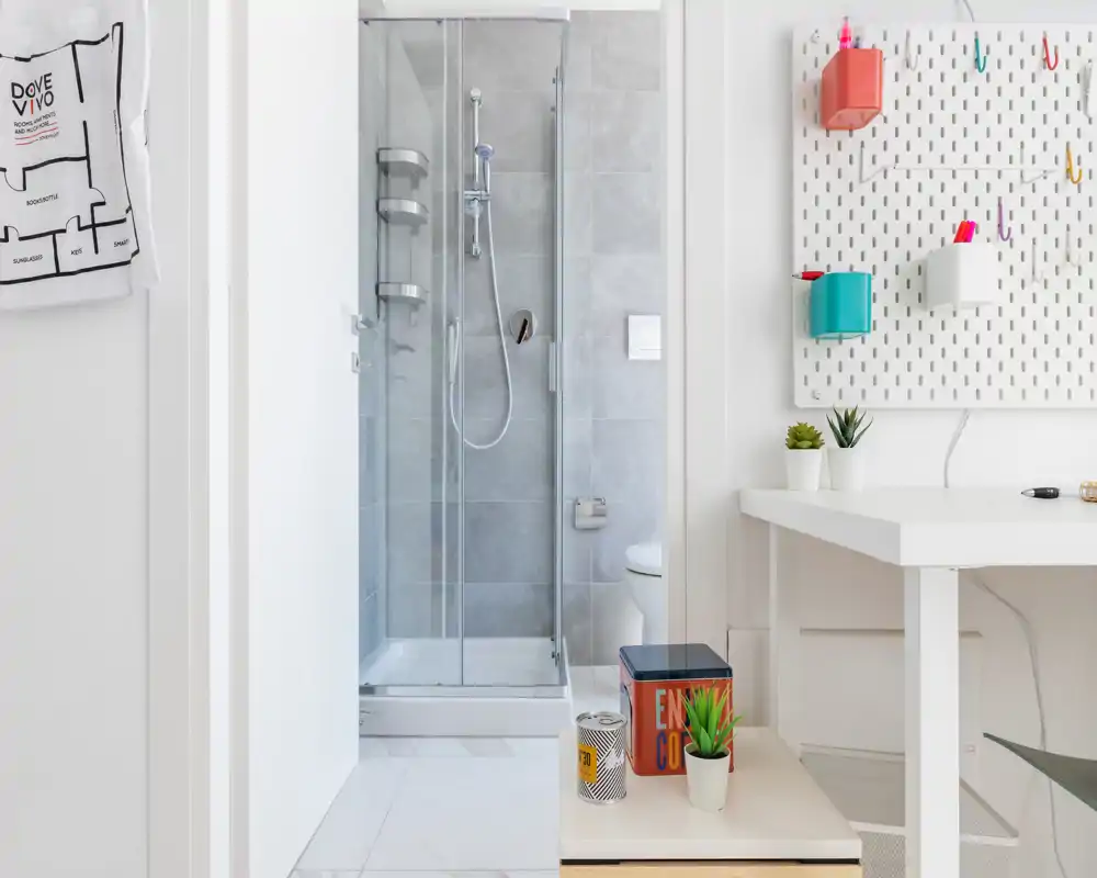 Small modern bathroom with a glass shower cubicle and neutral grey tiles, seen through a doorway from an adjacent workspace.
