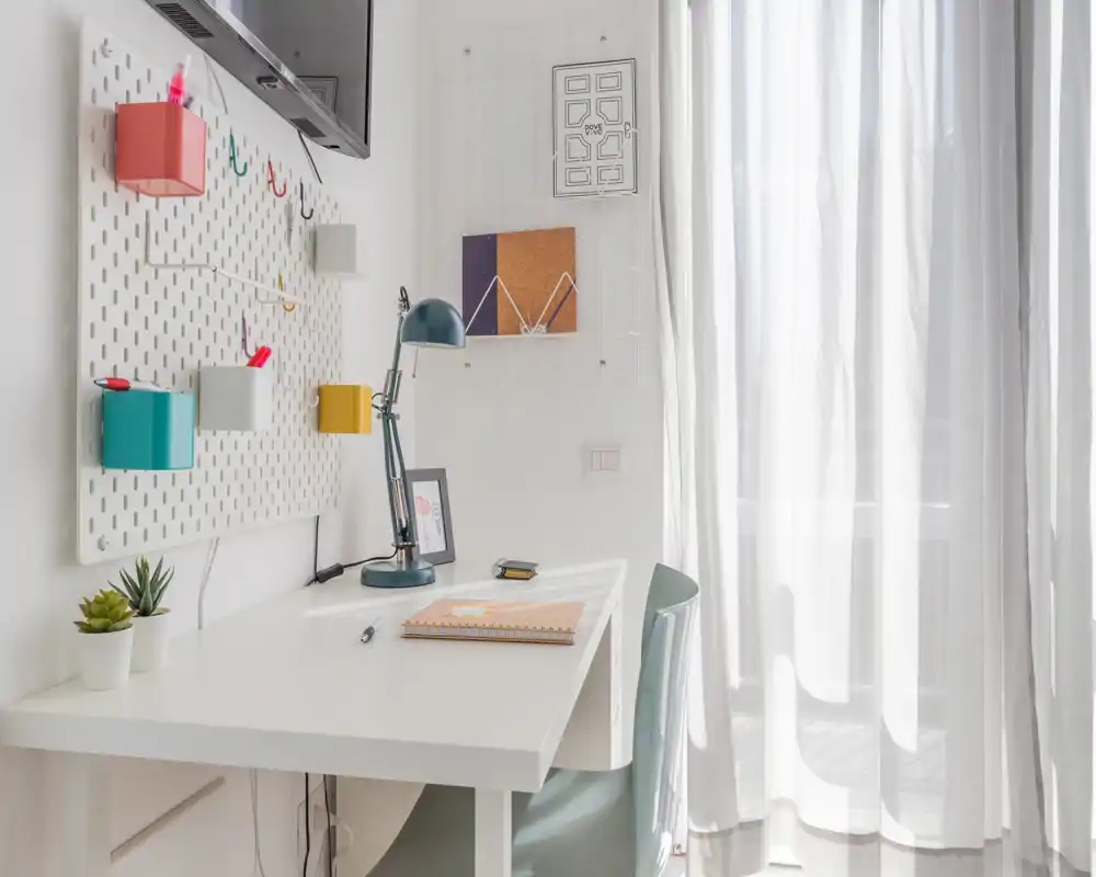 Bright, minimal desk area with a white worktable, lamp, small plants and wall organizers by sheer curtains letting in natural light.